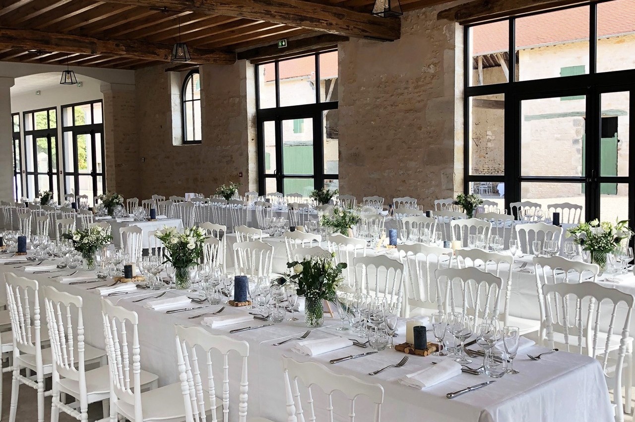 Salle de réception lumineuse avec de longues tables décorées de nappes blanches, fleurs et vaisselle élégante.