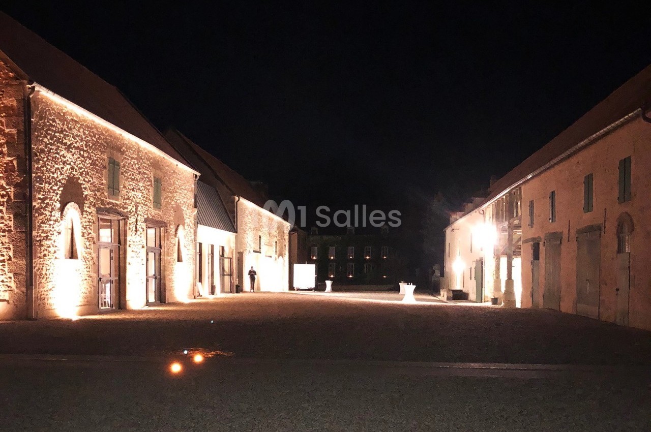 Cour intérieure éclairée la nuit, entourée de bâtiments en pierre avec des lumières soulignant leur architecture.