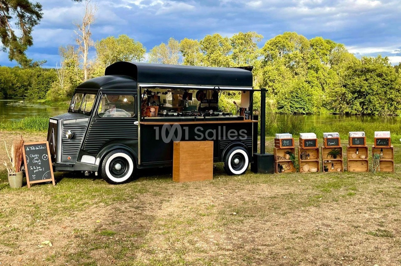 Camion noir aménagé en café ambulant, entouré de caisses en bois et situé dans un cadre naturel verdoyant.