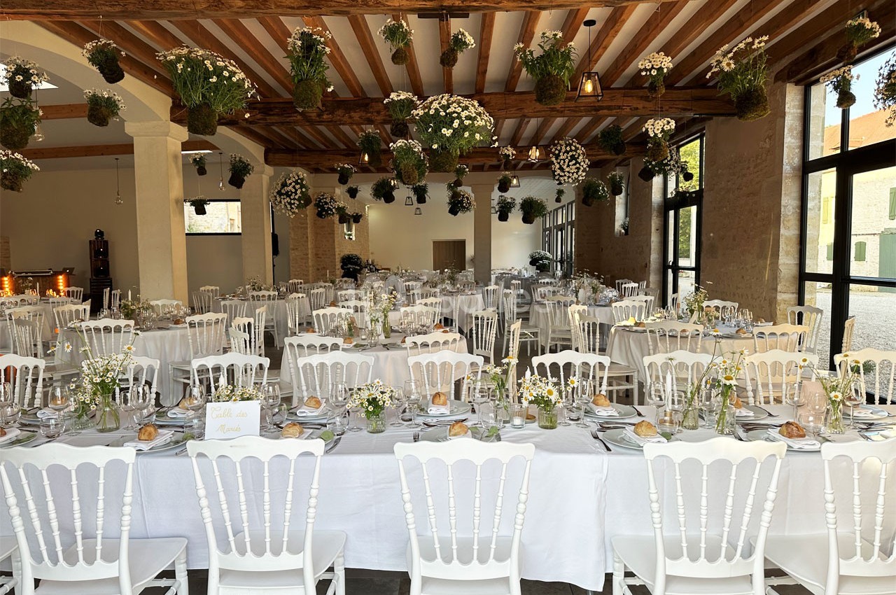 Salle de réception décorée avec des tables dressées, des chaises blanches et des suspensions florales au plafond.