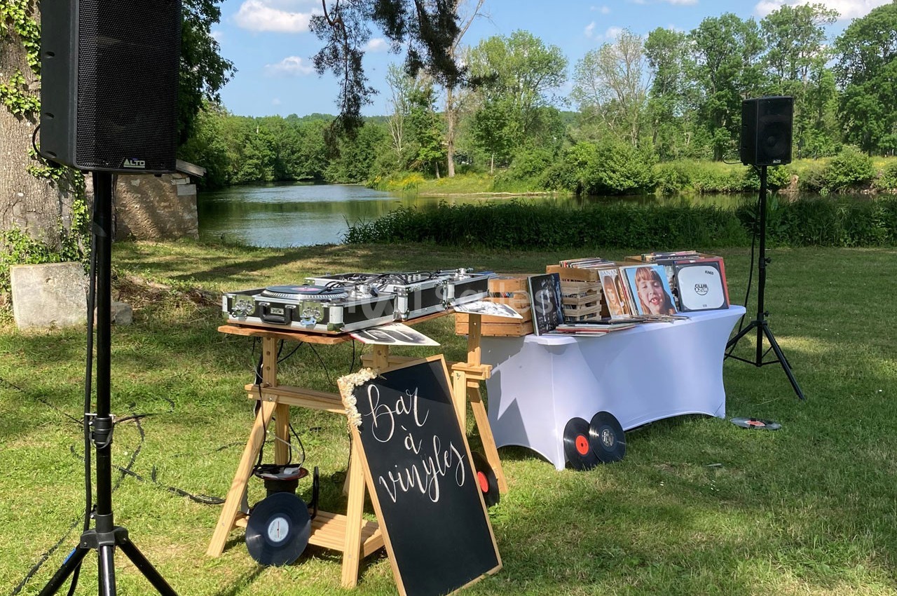 Table de mixage et vinyles installés en extérieur près d'un étang, entourés de verdure et d'enceintes.