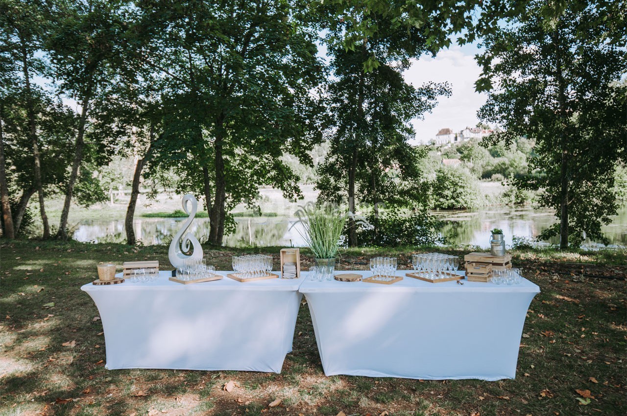 Deux tables blanches dressées en extérieur, entourées d'arbres, avec des décorations et des verres disposés.