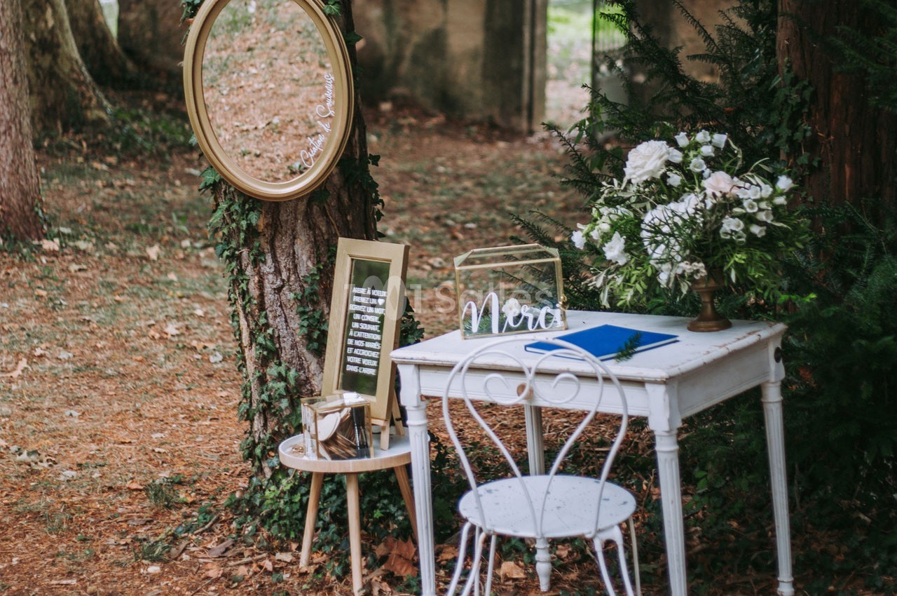 Table blanche décorée avec des fleurs, un miroir ovale fixé à un arbre et une chaise en métal dans un cadre boisé.