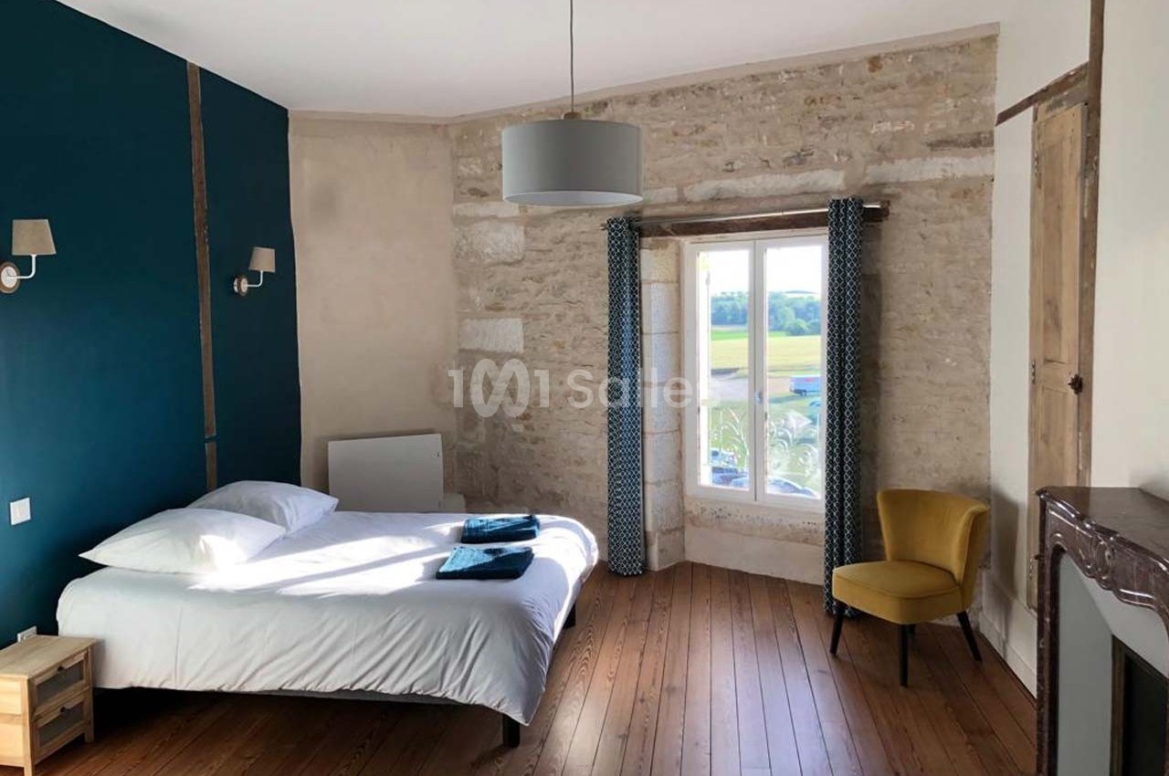 Chambre lumineuse avec un lit double, murs en pierre et peinture bleue, parquet en bois et fenêtre donnant sur la campagne.