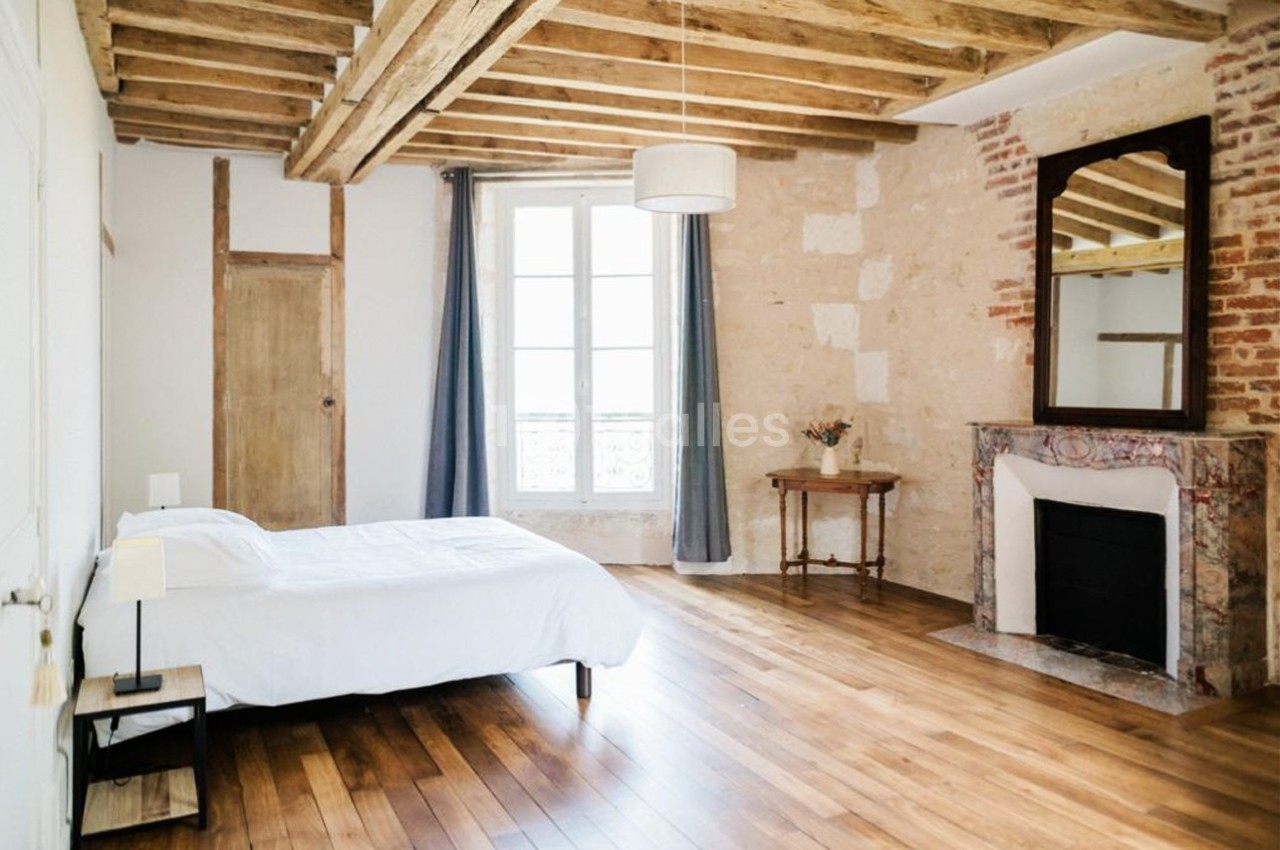 Chambre lumineuse avec lit simple, parquet en bois, cheminée décorative, murs en pierre et poutres apparentes.