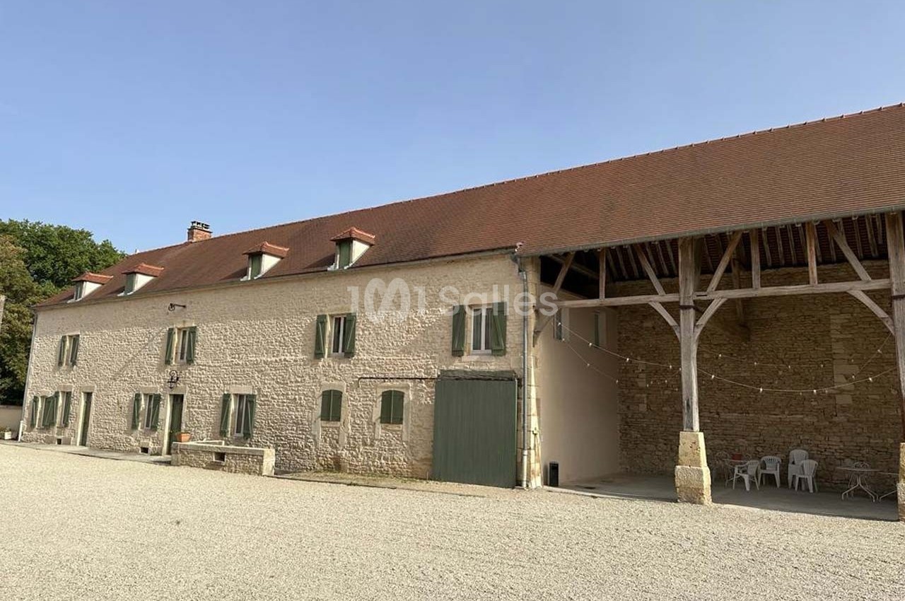 Bâtiment en pierre avec toit en tuiles, grandes fenêtres et une charpente ouverte, situé sur une cour gravillonnée.