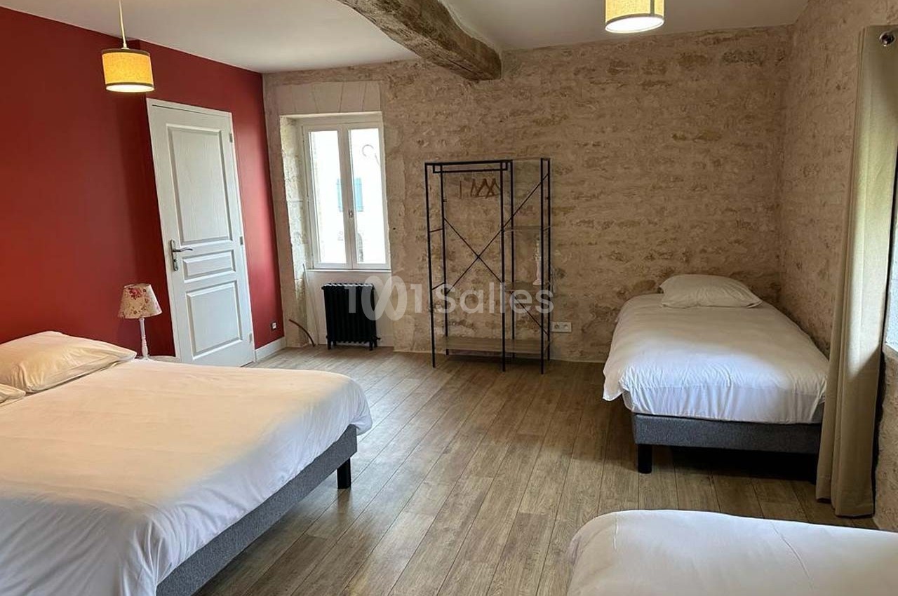Chambre avec trois lits simples, mur en pierre, parquet, mur rouge, et étagère métallique près d'une fenêtre.