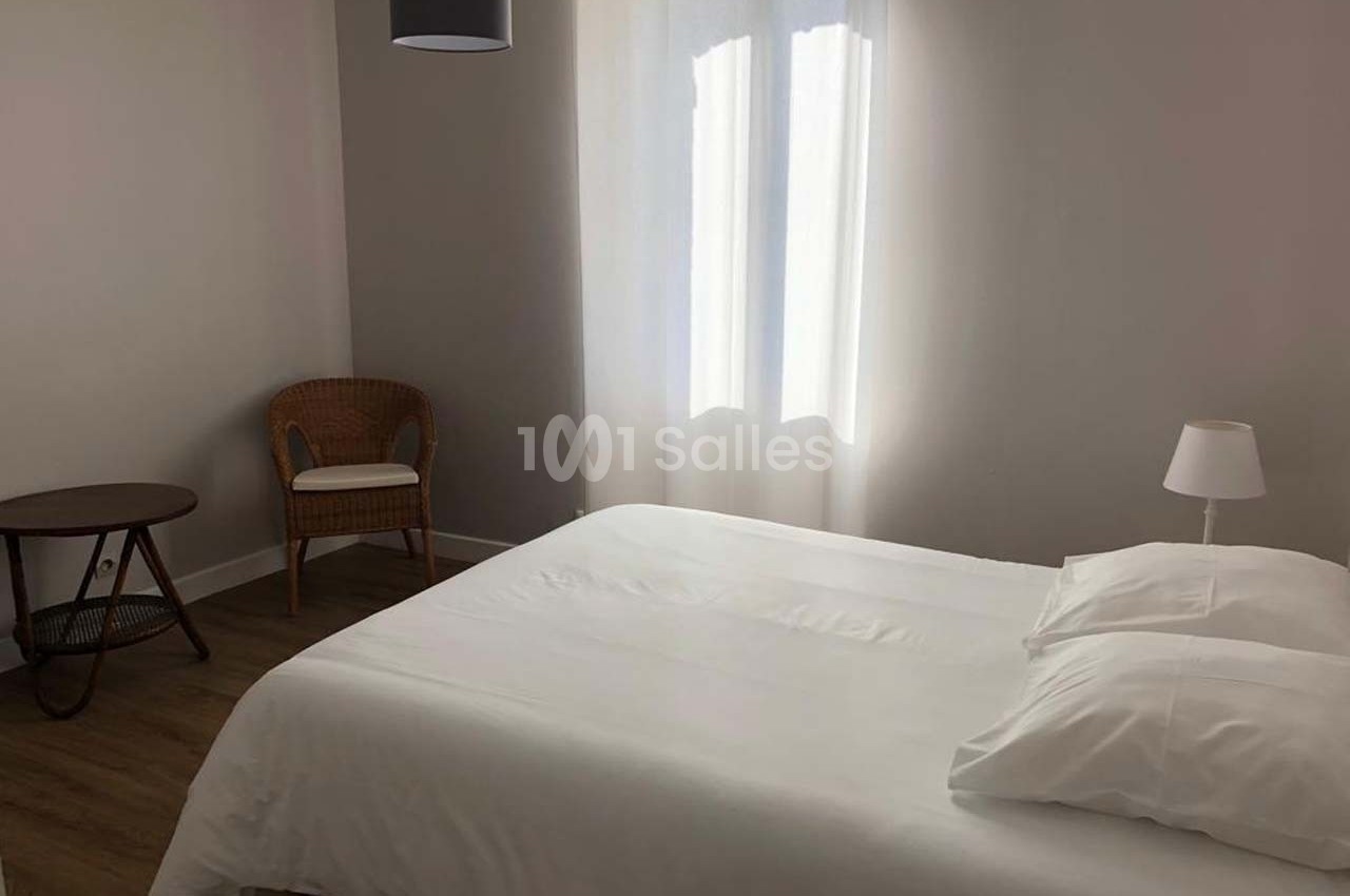Chambre lumineuse avec un lit double, une table ronde, une chaise en rotin et un lampadaire, sur un sol en parquet.