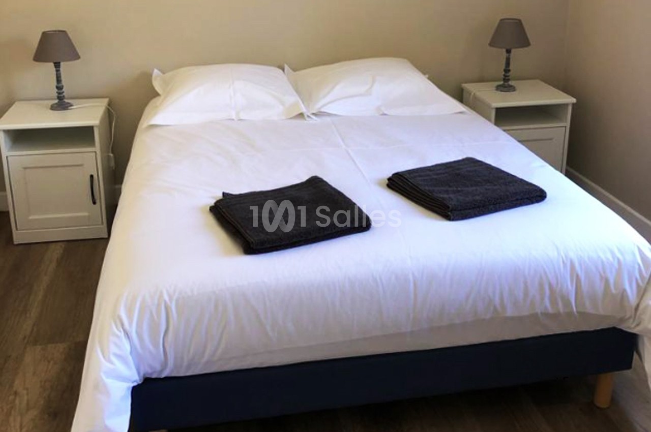 Lit double avec draps blancs, deux serviettes pliées au pied, tables de chevet avec lampes dans une chambre lumineuse.