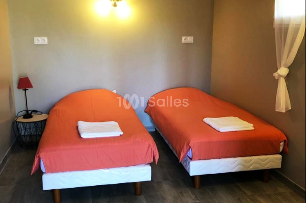 Deux lits simples avec draps orange et serviettes blanches, éclairés par une applique murale, dans une chambre sobre.