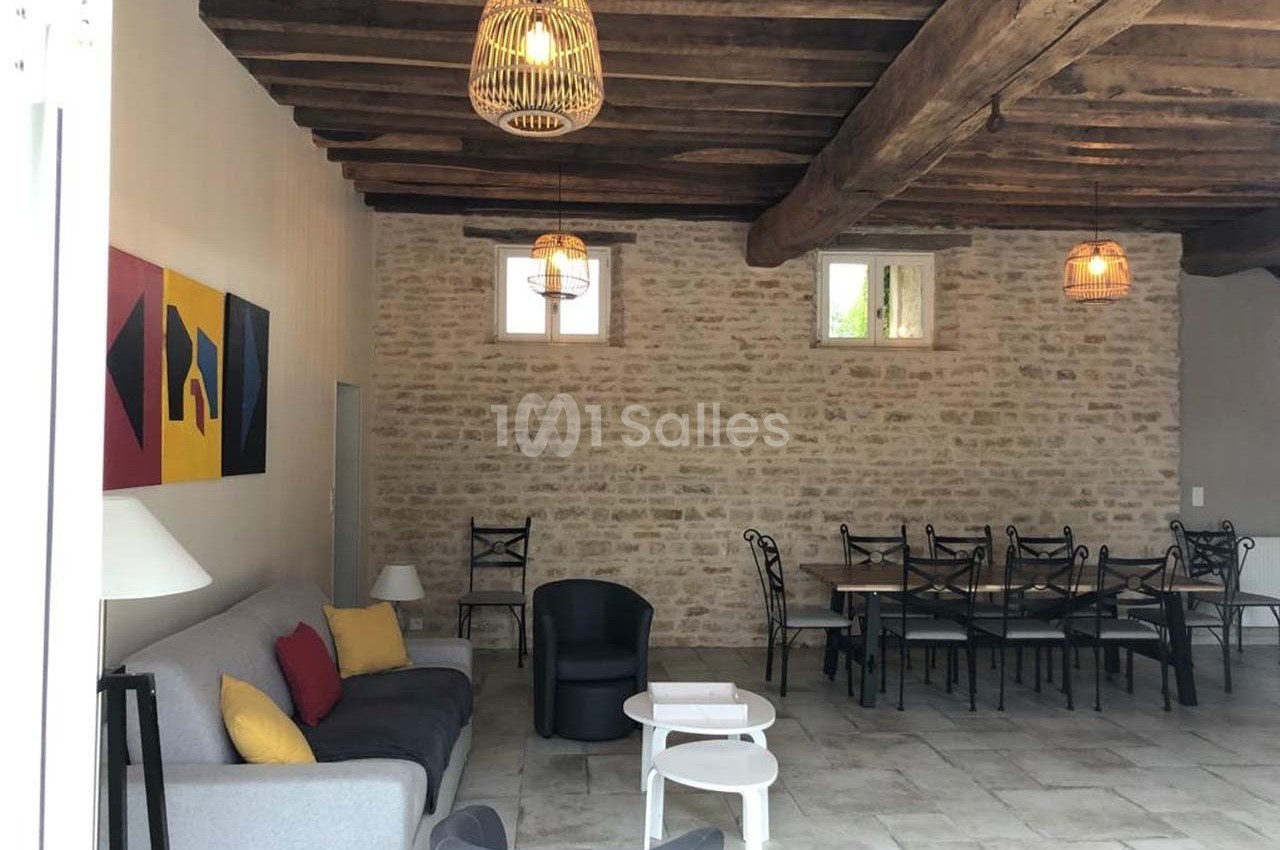 Salle de séjour avec murs en pierre, poutres apparentes, canapé gris, table à manger et chaises noires.