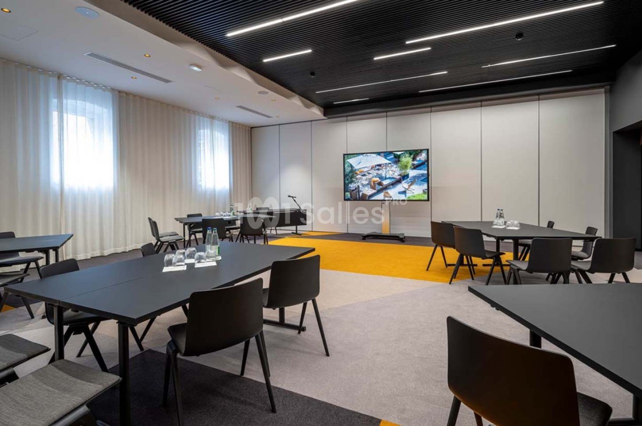 Salle de réunion moderne avec tables noires, chaises, écran large et éclairage naturel provenant de fenêtres.