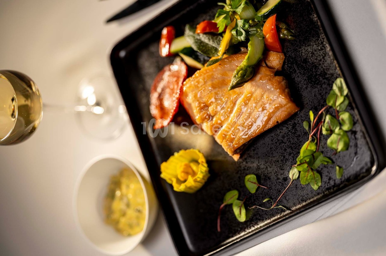 Assiette gastronomique avec filet de poisson grillé, légumes colorés, purée décorative et verre de vin en arrière-plan.