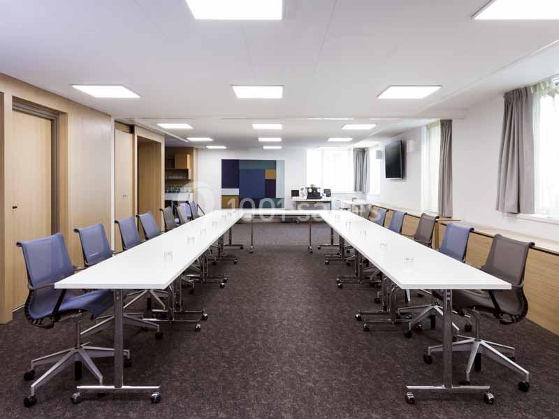Salle de réunion lumineuse avec deux rangées de tables blanches et chaises, équipée d'un écran et d'un tableau.