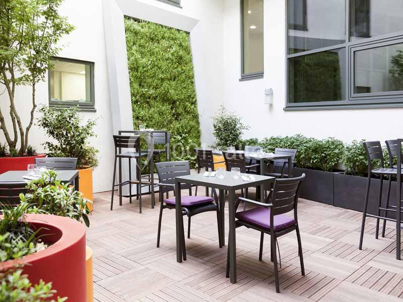 Terrasse extérieure avec tables et chaises entourées de plantes et d'un mur végétal, dans un cadre lumineux.