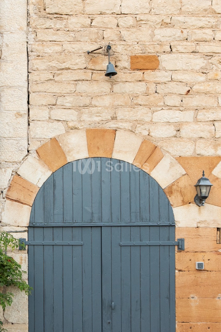 Porte en bois bleu arrondie encadrée de pierres, avec une lampe et une cloche fixées sur un mur en pierre beige.