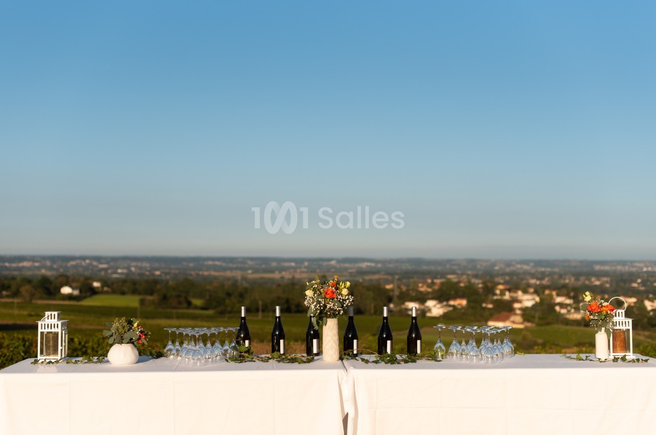 Deux tables blanches dressées avec des bouteilles de vin, des verres et des décorations florales, surplombant un paysage…