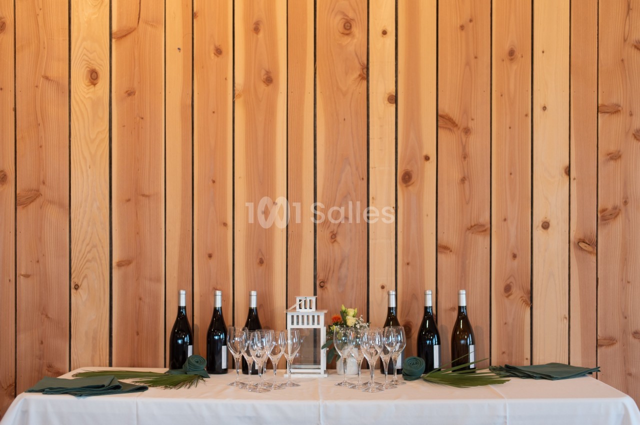 Table dressée avec des verres à vin, des bouteilles et des serviettes vertes, devant un mur en bois clair.