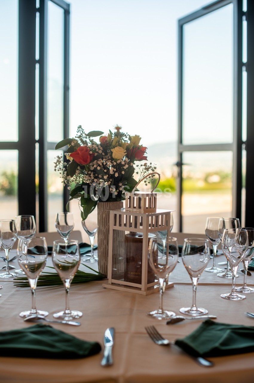 Table dressée avec des verres, des serviettes vertes, un centre de table floral et une lanterne, devant des portes ouvertes.