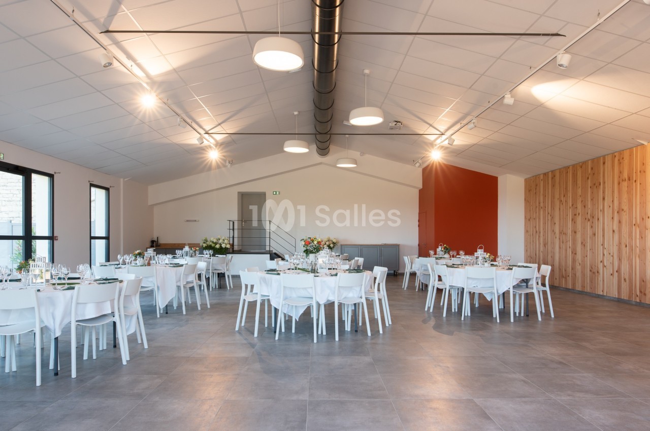 Salle de réception lumineuse avec tables rondes dressées, décorée de fleurs, et murs aux tons clairs et bois.