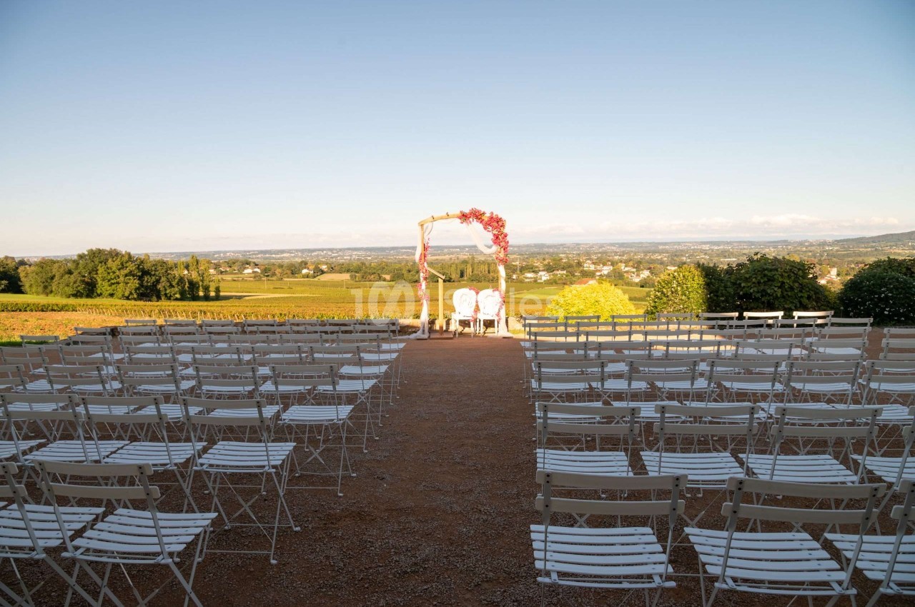 Chaises blanches alignées en extérieur face à une arche décorée de fleurs, avec vue sur un paysage de campagne.