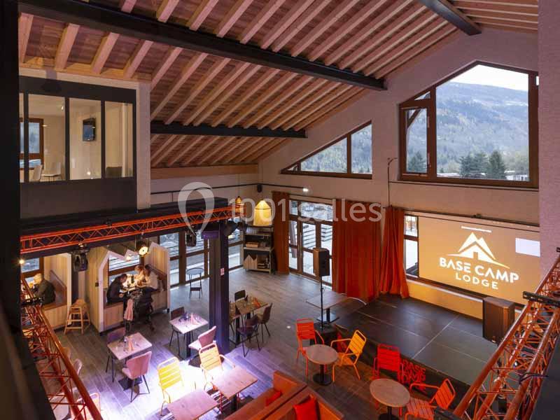 Salle lumineuse avec poutres apparentes, grandes baies vitrées, mobilier coloré et écran affichant ’Base Camp Lodge’.