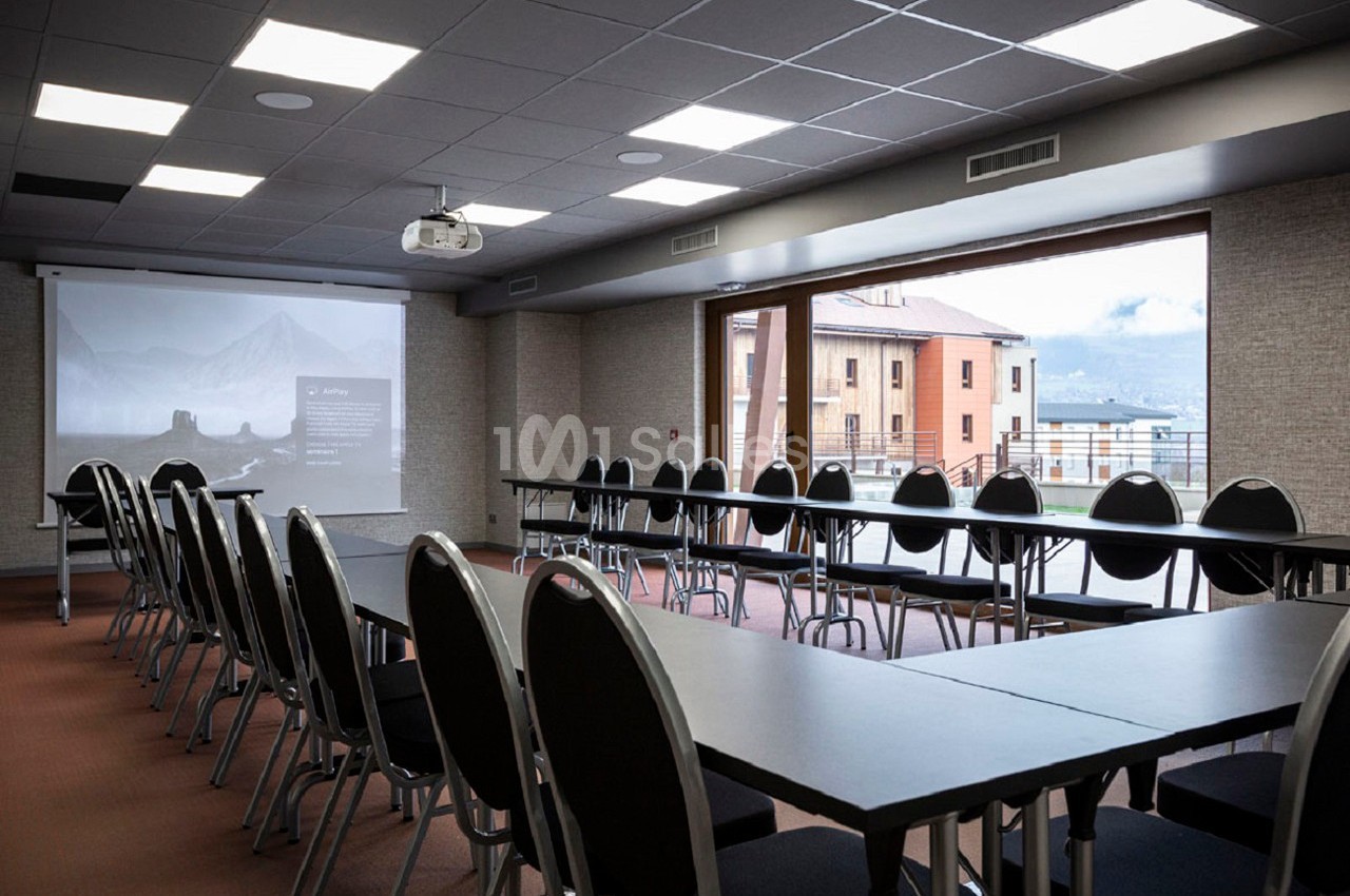 Salle de réunion lumineuse avec tables et chaises alignées, écran de projection et vue sur des bâtiments extérieurs.