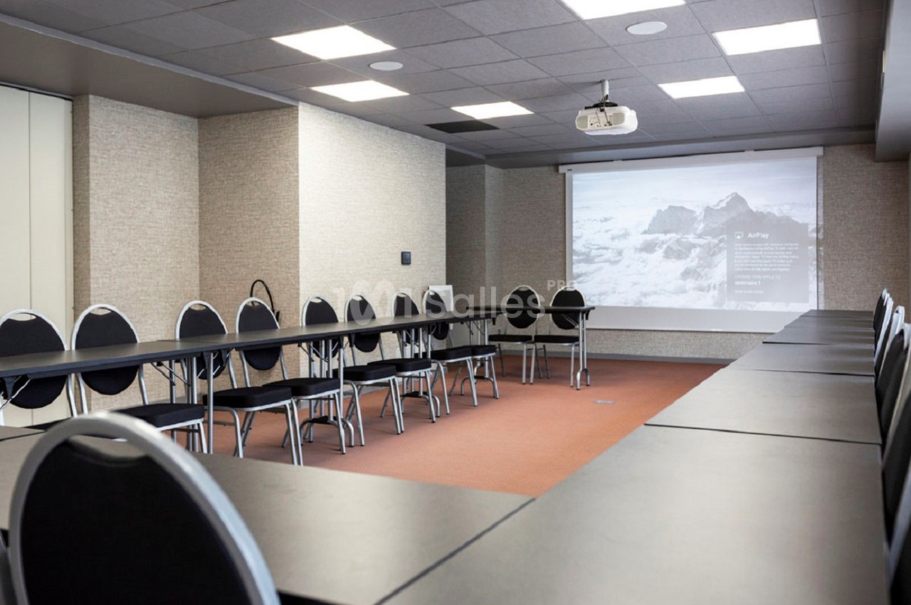Salle de réunion équipée de tables en U, chaises noires, projecteur et écran affichant une image de montagne.