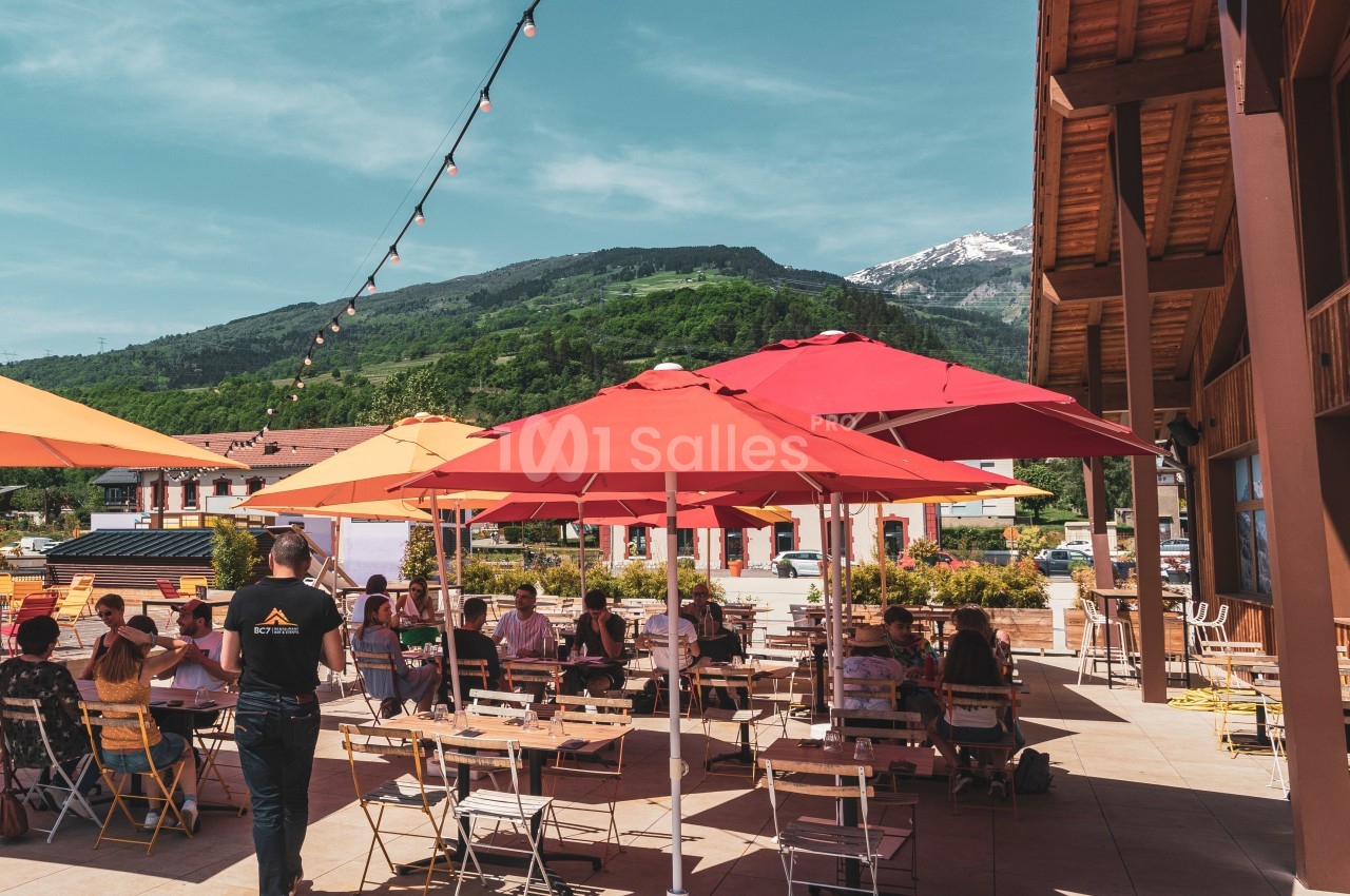 Terrasse ensoleillée avec des tables et chaises sous des parasols colorés, entourée de montagnes verdoyantes.