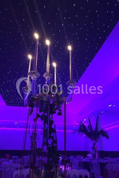 Lustre à bougies allumées dans une salle éclairée en violet, avec plafond étoilé et décorations végétales.