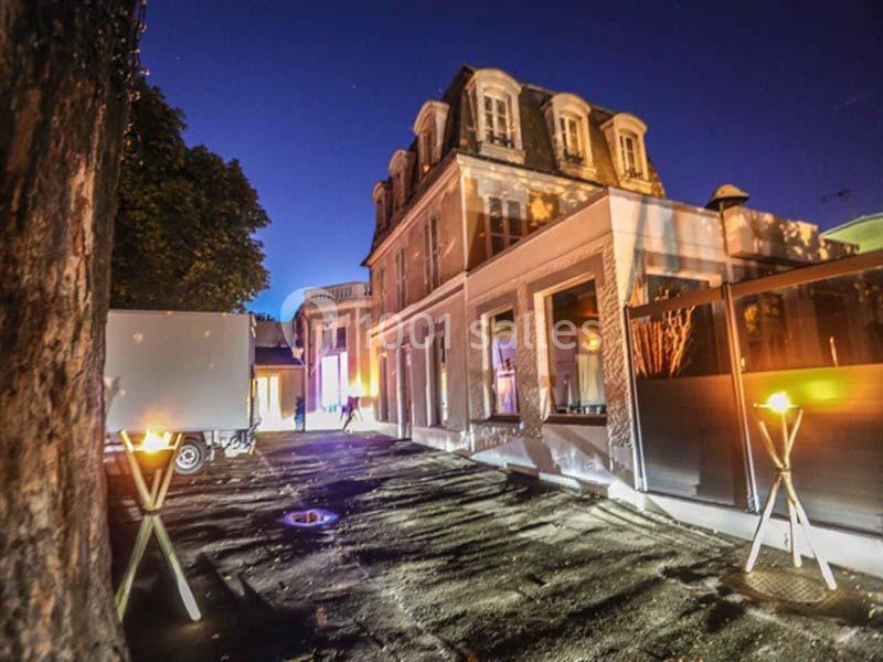 Façade d'un bâtiment ancien éclairé de nuit, avec des torches allumées et un arbre au premier plan.