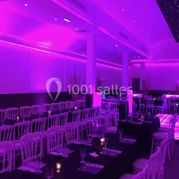 Salle de réception éclairée par une lumière violette, avec des tables dressées et des chaises blanches alignées.