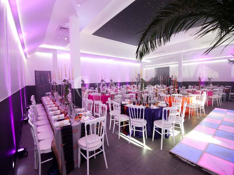 Salle de réception décorée avec des tables dressées, des chaises blanches et un éclairage violet tamisé.
