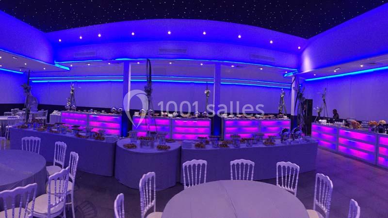 Salle de réception illuminée de lumières violettes et bleues, avec tables dressées et buffet en arrière-plan.