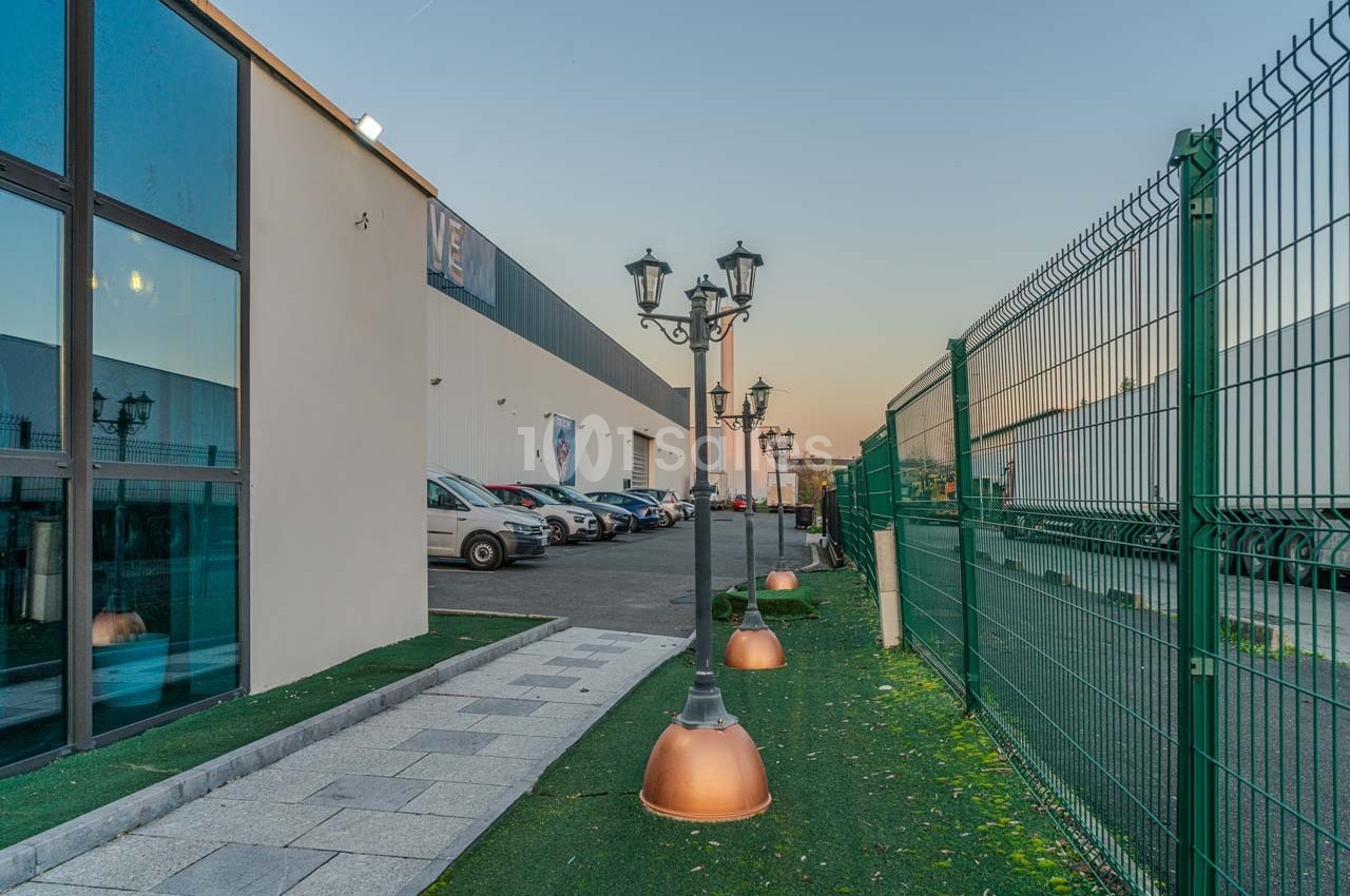 Allée bordée de lampadaires et clôturée, menant à un bâtiment industriel avec des voitures stationnées.