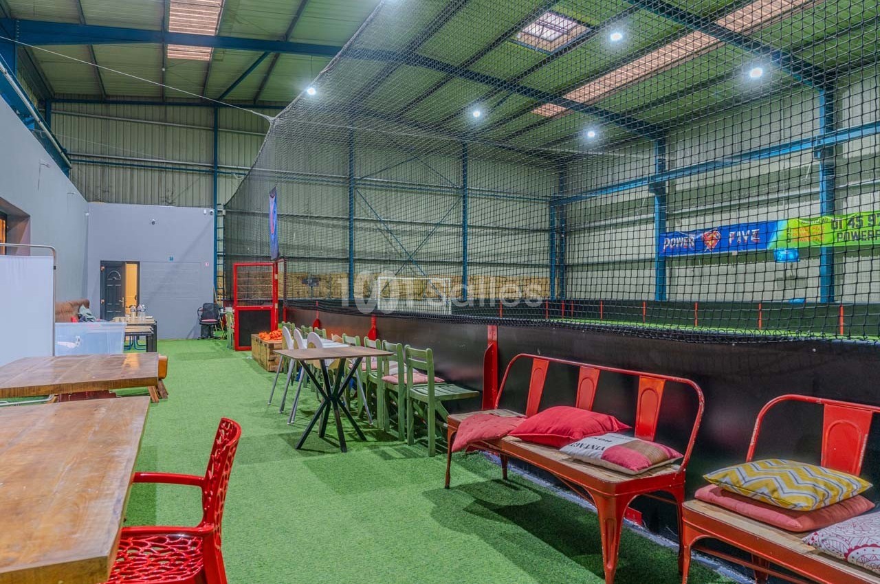 Salle intérieure avec terrain de sport entouré de filets, tables et chaises disposées sur un sol en gazon synthétique.