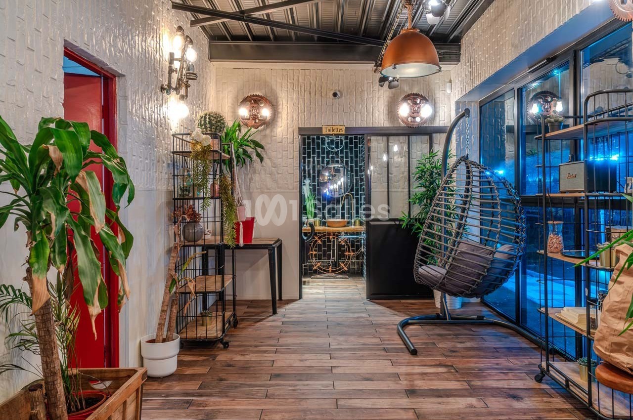 Couloir décoré de plantes, meubles en bois et luminaires modernes, menant à une pièce avec une table et des chaises.
