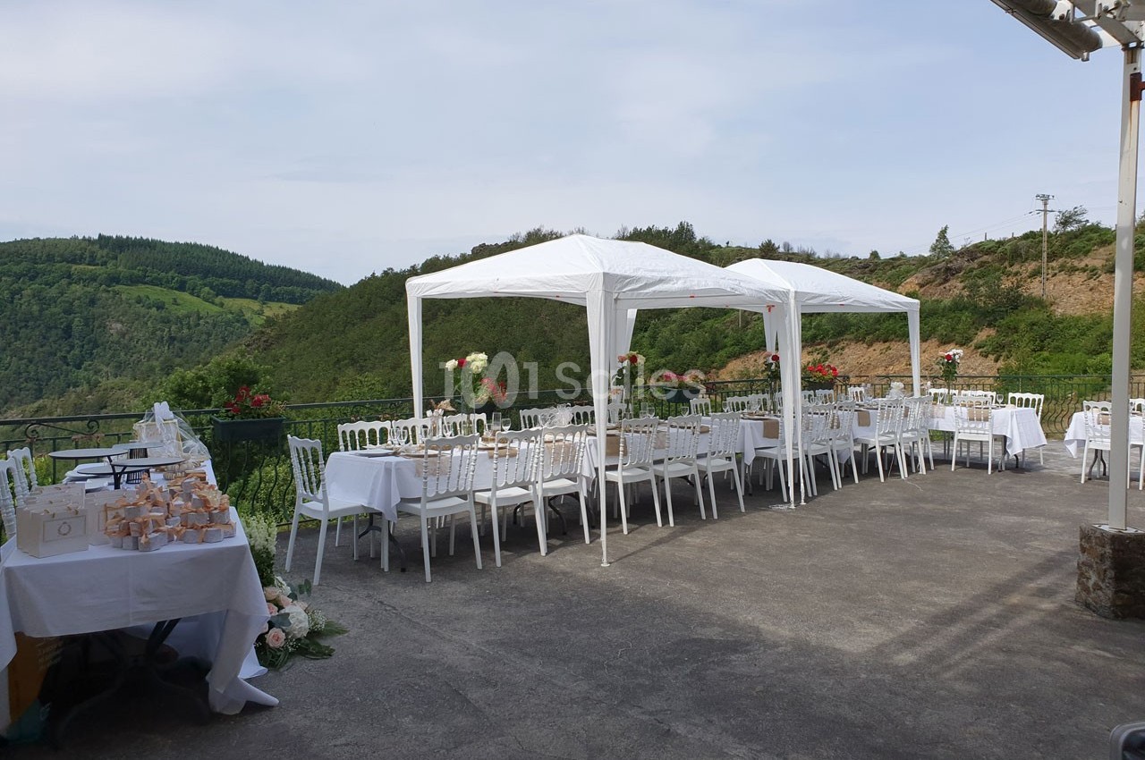 Tables et chaises blanches disposées sous des tonnelles sur une terrasse avec vue sur des collines verdoyantes.