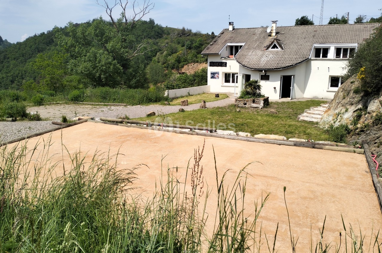 Terrain de pétanque en sable devant une maison blanche entourée de verdure et de collines.