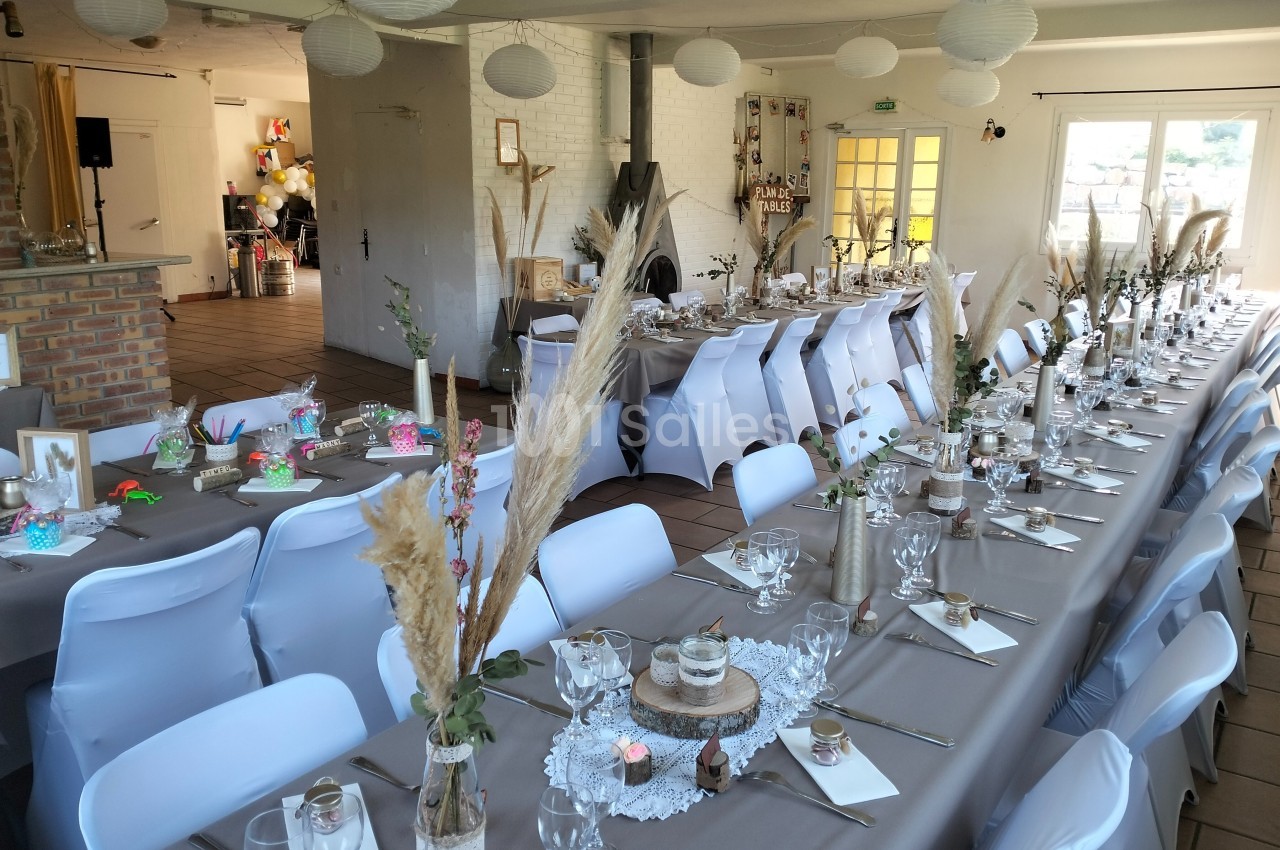 Salle décorée pour un événement avec longues tables dressées, nappes grises, chaises blanches et ornements naturels.