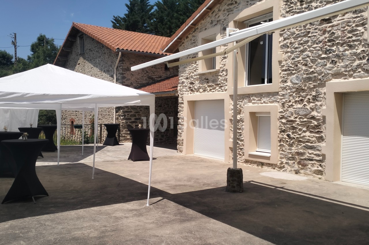 Cour extérieure avec des bâtiments en pierre, un barnum blanc et des tables hautes noires sur une terrasse ensoleillée.