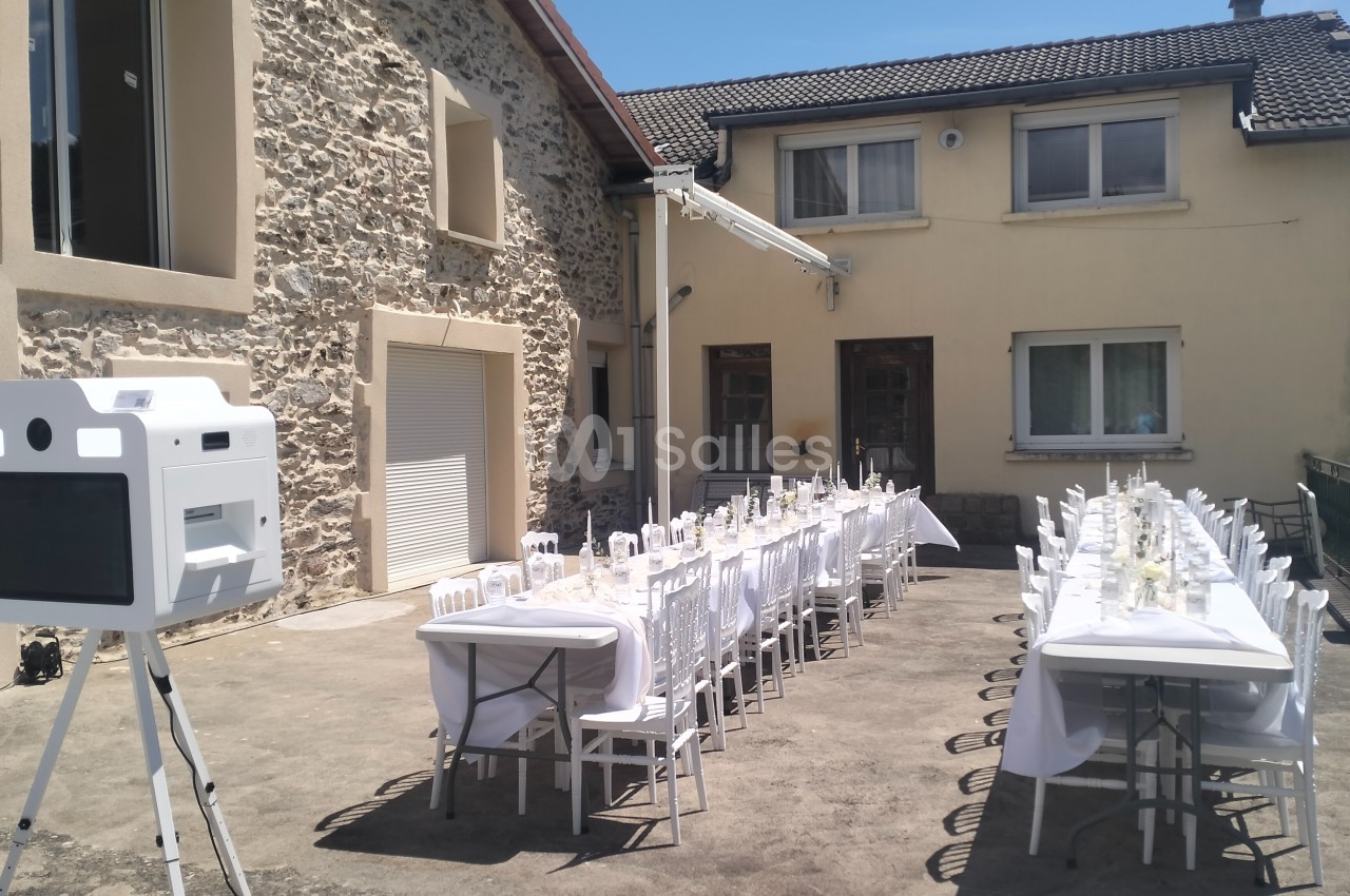 Tables dressées pour un repas en extérieur devant une maison en pierre et crépi, avec une borne photo à gauche.