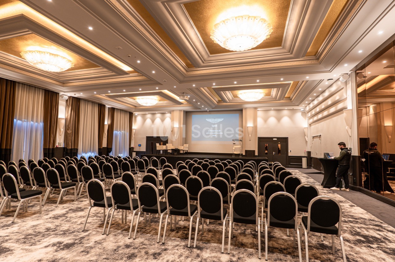 Salle de conférence élégante avec rangées de chaises noires, écran de projection allumé et éclairage raffiné.