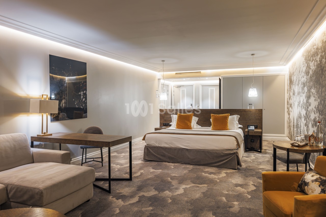 Chambre d'hôtel avec lit king-size, zone salon et bureau, éclairage indirect, mobilier bois et tons neutres.