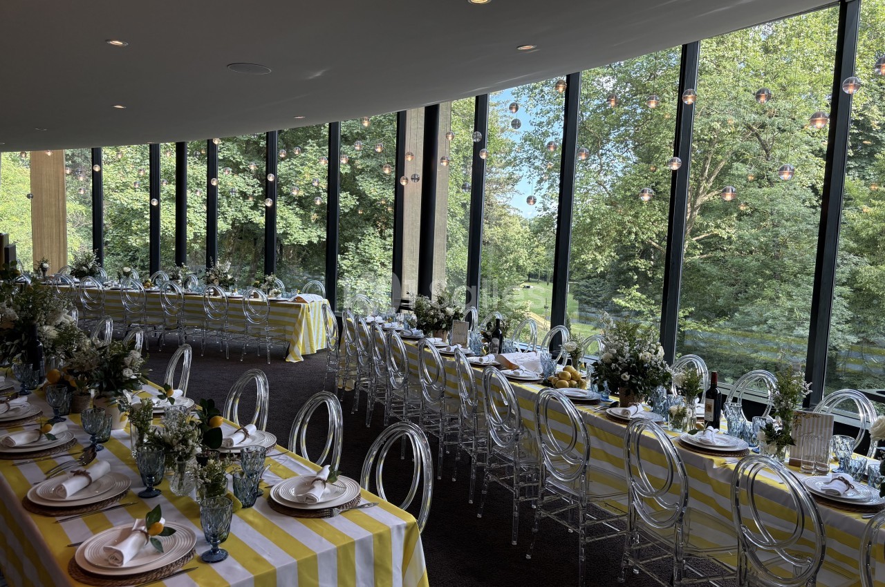 Salle dressée pour réception avec tables aux nappes jaune et blanc, chaises transparentes, vue panoramique sur parc boisé.