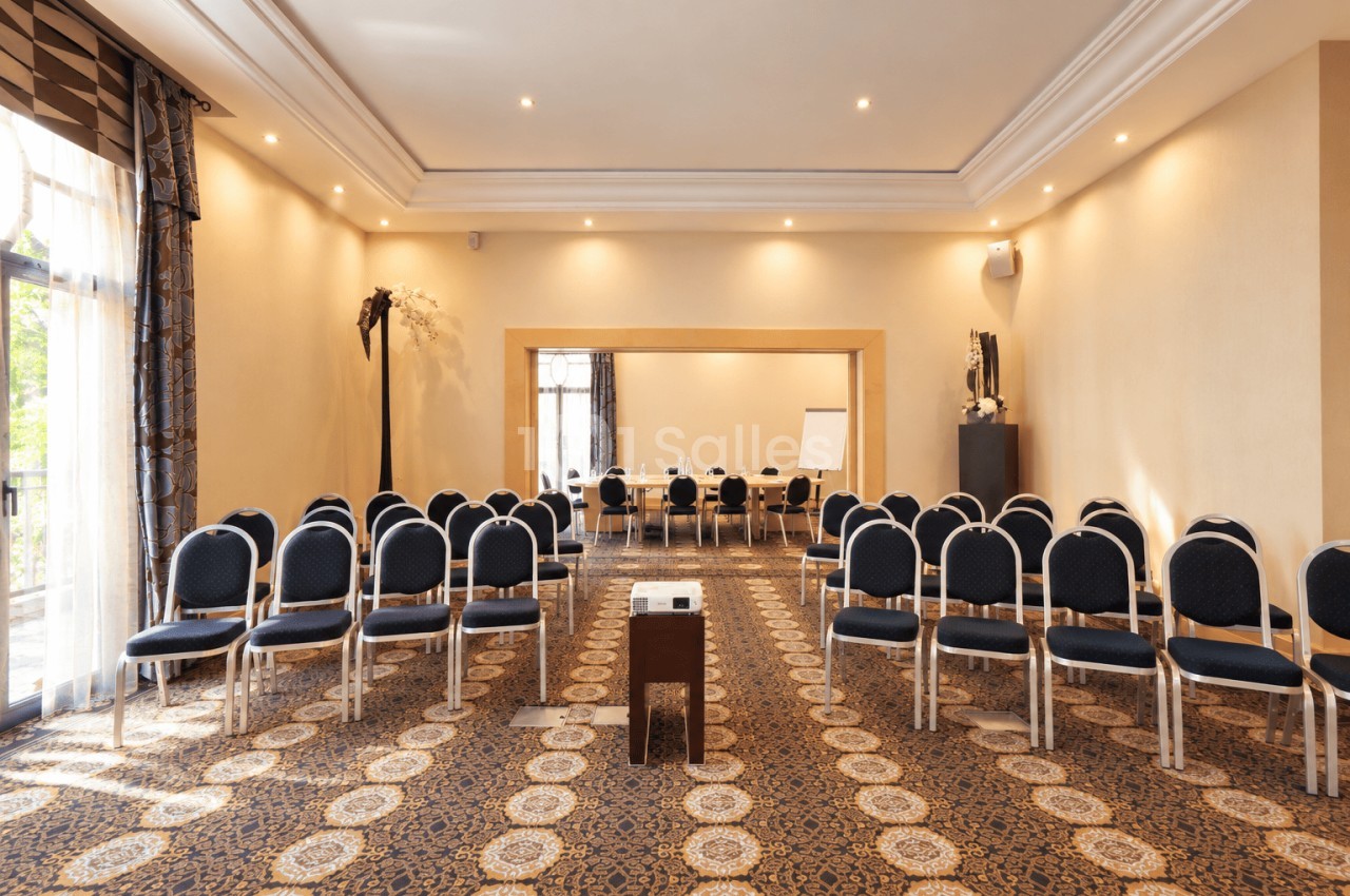 Salle de conférence beige avec chaises en demi-cercle face à un pupitre, accès à une salle attenante.
