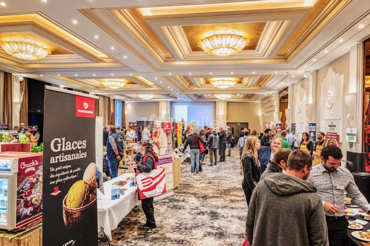 Salle de réception aménagée pour un salon professionnel avec stands de prestataires, plafond caisson doré et cristalliers, environ cent cinquante visiteurs en circulation.