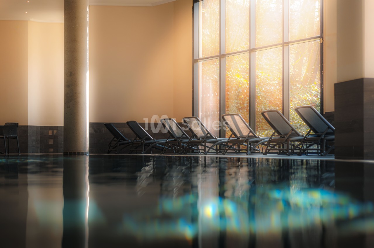 Espace aquatique intérieur avec chaises longues alignées face à une baie vitrée donnant sur un jardin automnal.