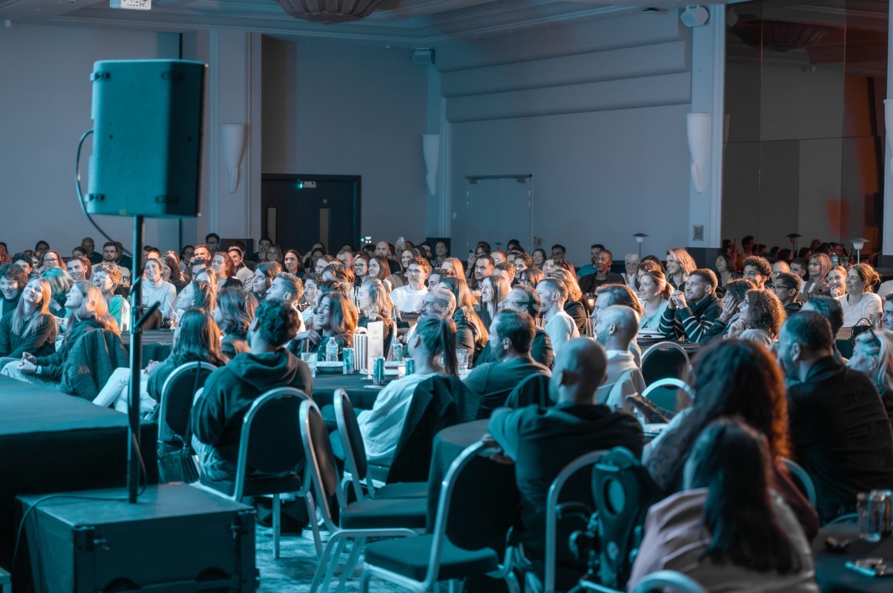Des personnes assises dans une salle de conférence, attentives à une présentation sur une scène.