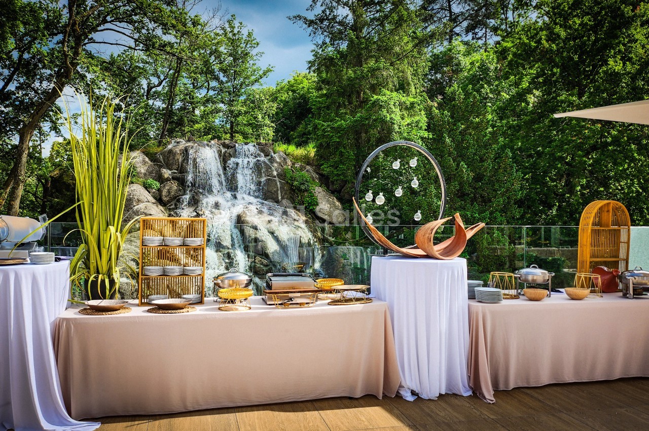 Buffet en plein air avec nappes blanches, plats variés et une cascade entourée de verdure en arrière-plan.